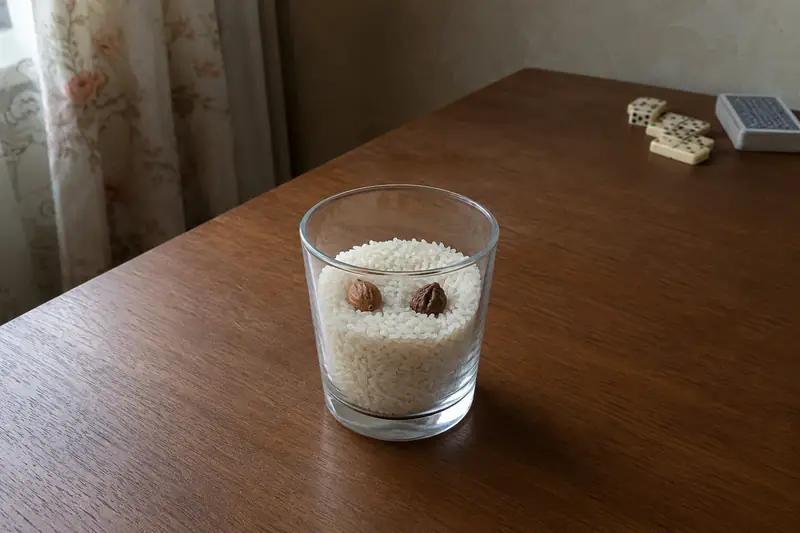 Стеклянный стакан с сухим рисом и одной гладкой абрикосовой косточкой и одной тёмной сливовой косточкой на простом деревянном столе в тихом свете, поодаль — обычное игральное домино и колода карт убраны в сторону — чего при гадании на косточках не делают