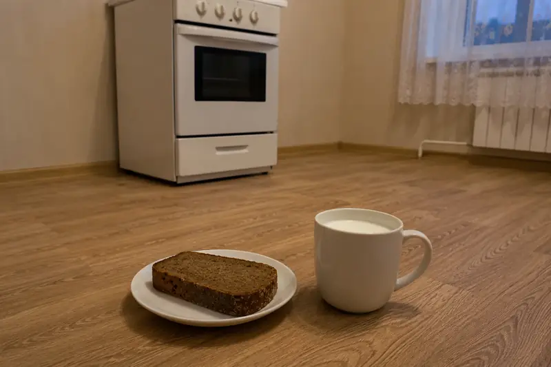 Небольшое простое блюдце с куском чёрного ржаного хлеба и обычная белая чашка с молоком на полу у плиты в пустой новой кухне при тёплом вечернем свете — новоселье и угощение домовому