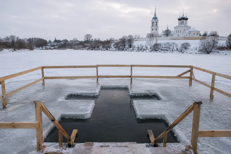 Прорубь-иордань в форме креста на льду замёрзшей реки с деревянными ограждениями у зимнего православного храма — купание в проруби на Крещение