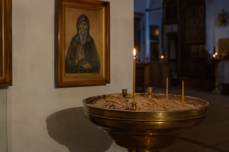 Тонкая восковая свеча горит перед иконой святого в храме, рядом подсвечник с песком, спокойный приглушённый свет — молебен и свеча чудотворцу в храме