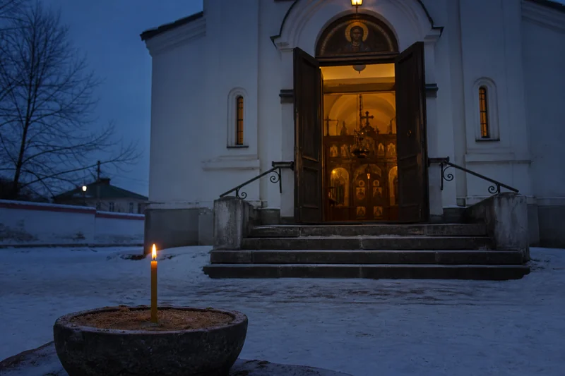 Вход небольшого православного храма с мягким вечерним светом и одной свечой на переднем плане — богослужение Рождества Христова