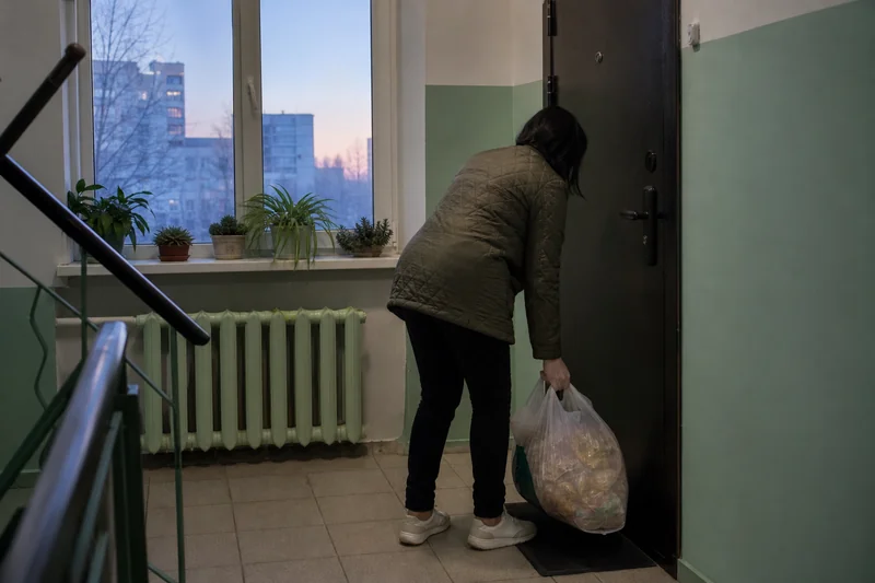Женщина в куртке берёт сумку с товаром у входной двери на рассвете, за окном светлеющее утреннее небо — когда читать заговор на быструю продажу